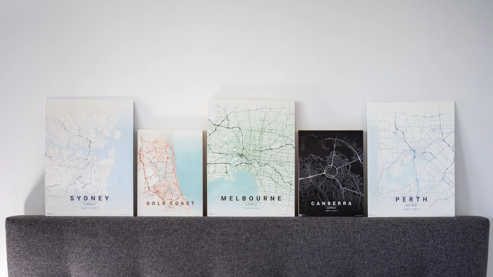 Wood-board map prints of Sydney, Gold Coast, Melbourne, Canberra, and Perth leaning on a headboard